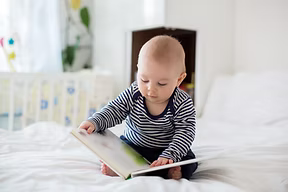 Kleinkind sitzt auf dem Bett und schaut in ein Buch – Symbol für den individuellen Schritt-für-Schritt Plan im Online Schlafcoaching.